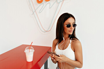 Close-up of image of pretty caucasian young girl in sunglasses listening music in headphones and using smartphone. Brunette with straight hair sitting in stylish dressed in white top.