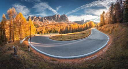 Winding road in mountains and orange trees at colorful sunset in autumn in Italy. Landscape with forest, beautiful roadway, rocks, blue sky in fall. Highway, woods with orange leaves in Alps. Nature