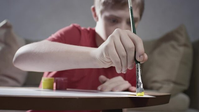 Portrait of a boy with Down Syndrome. Cheerful kid draws with paints and brush at home. Creates art and painting. A person with special needs. Chromosomal genetic disorder in a child.