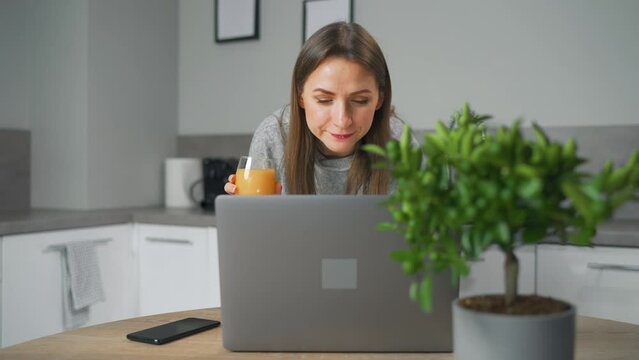 Woman is standing in the kitchen, drinking juice and working on a laptop, doing remote work, texting and resolving issues
