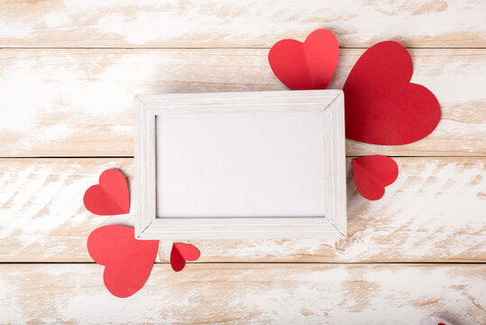 Photo frame between Valentines day romantic decorating with paper hearts on a white wooden table. Top view, copy space.