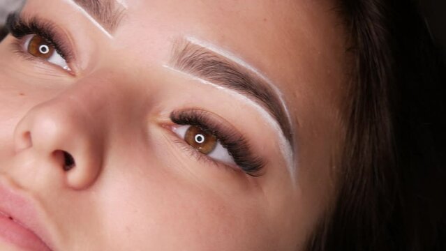 Master applies white markings to correct shape of eyebrows with a special brush. The beautiful face of young girl model on sofa in a beauty salon before microblading, permanent makeup eyebrow tattoo