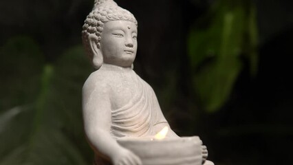 Buddha made of stone on wooden table surface at natural exotic jungle green background with monstera plant. Buddha statue with burning candle fire and water steam.