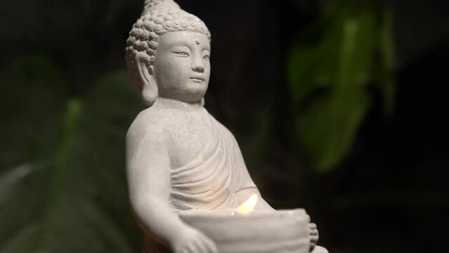 Buddha made of stone on wooden table surface at natural exotic jungle green background with monstera plant. Buddha statue with burning candle fire and water steam.