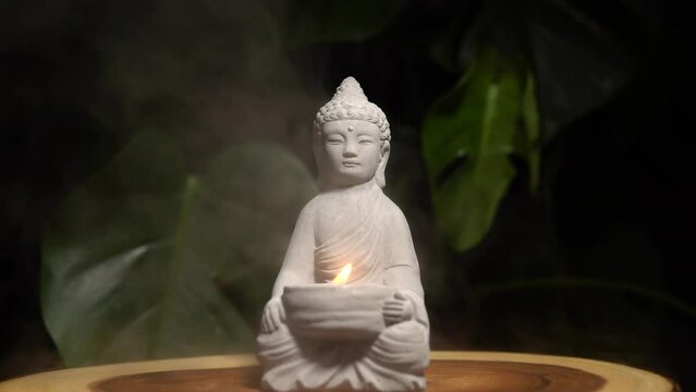 Buddha statue with burning candle fire and water steam. Buddha made of stone on wooden table surface at natural exotic jungle green background with monstera plant.