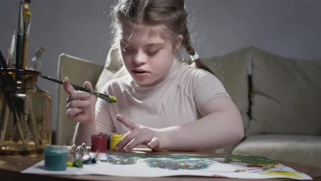 Portrait of a girl with Down Syndrome. The little girl draws with paints and a brush at home. Creates art and creativity. A person with special needs. Chromosomal genetic disorder in a child.