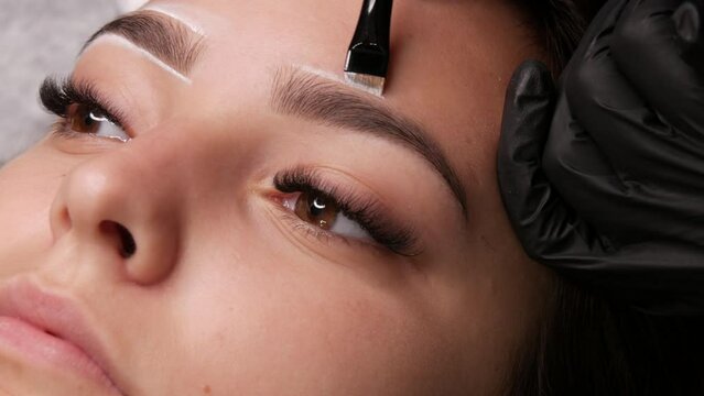 Master applies white markings to correct shape of eyebrows with a special brush. The beautiful face of young girl model on sofa in a beauty salon before microblading, permanent makeup eyebrow tattoo