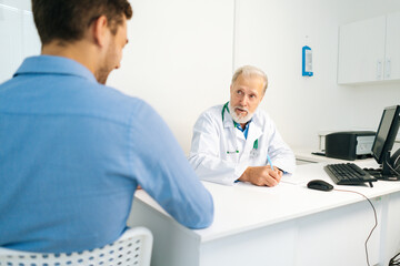 Fototapeta na wymiar Mature male adult doctor talking with unrecognizable young man patient during medical visit sitting at table in medical office. View from back to older senior physician consulting man at hospital.