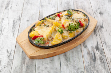 Pollock with vegetables. Baked with cheese sauce and cream. In a frying pan on a stand. Light wooden background. View from above.