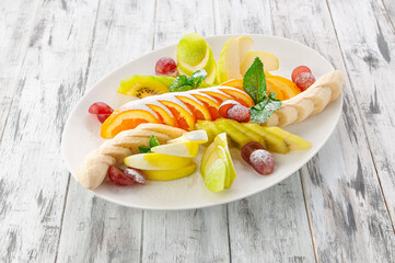 A set of snacks from fruits cut into slices. Decorated with powdered sugar. In a white plate. Light wooden background. View from above.