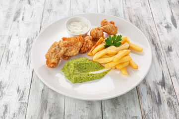 Pollock fillet fried in batter with french fries. Green pea puree with garlic sauce. On a white plate. Light wooden background. View from above.