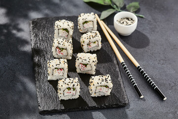 Maki sushi on dark slate. California maki with sesame. Sushi roll with crab, cucumber inside, sesame outside. Style concept japanese menu with black background, leaves and hard shadow.