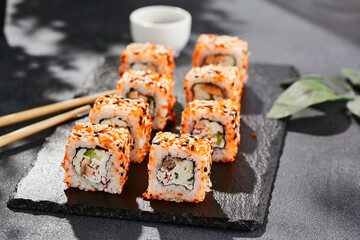 Maki sushi on dark slate. California maki with masago and sesame. Sushi roll with crab, cucumber inside, tobiko outside. Style concept japanese menu with black background, leaves and hard shadow.