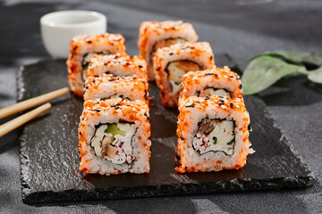 Maki sushi on dark slate. California maki with masago and sesame. Sushi roll with crab, cucumber inside, tobiko outside. Style concept japanese menu with black background, leaves and hard shadow.