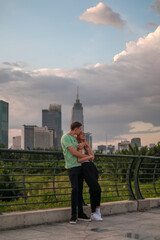 Lovely mixed couple standing on the bridge in the park in metropolis and cuddling. Love people and happiness concept. Skyscrapers on the background