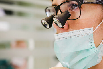 Young female doctor wearing dental loupe binoculars and medical mask at her work in dental clinic office.