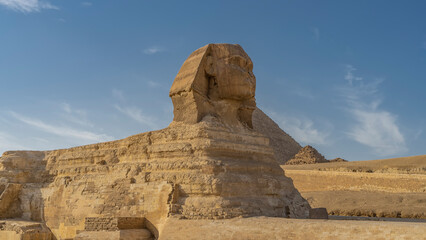 Fototapeta na wymiar Giant statue of the Great Sphinx against the blue sky. Profile view. The stone layered structure of the sculpture is visible. In the distance are the pyramids of Menkaure. Egypt. Giza