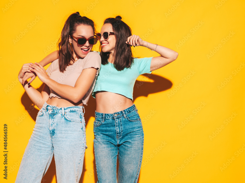 Wall mural Two young beautiful smiling blond hipster female in trendy summer clothes. Sexy carefree women posing near yellow wall in studio. Positive models having fun. Cheerful and happy. In sunglasses