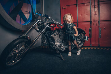 Fototapeta na wymiar a girl in a leather black dress stands on the background of a motorcycle red garage and a huge fan holding the handle of the bike with her hand
