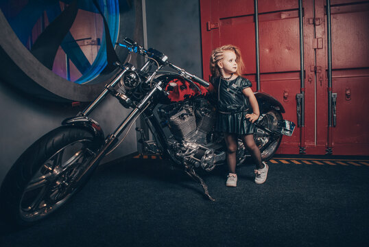 a girl in a leather black dress stands on the background of a motorcycle red garage and a huge fan holding the handle of the bike with her hand