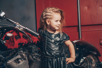 Plakat a little charming girl stylish in chains and leather latier stands against the background of a bike and a red iron garage