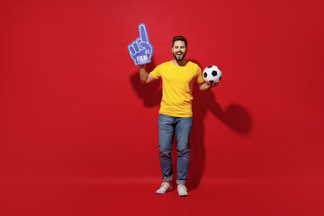 Full size body length young bearded man football fan in yellow t-shirt cheer up support favorite team hold soccer ball fan foam glove finger up isolated on plain dark red background studio portrait.