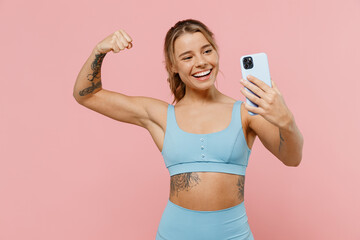 Young strong sporty athletic fitness trainer woman wear blue tracksuit spend time in home gym do selfie shot on mobile cell phone show muscles isolated on plain pink background. Workout sport concept.