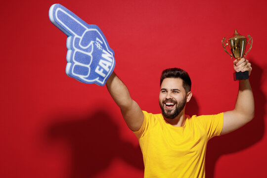 Smiling fun young bearded man 20s in yellow t-shirt cheer up support favorite sport team look aside hold fan foam glove finger up champion cup isolated on plain dark red background studio portrait