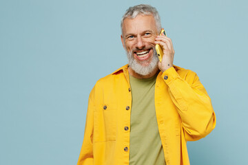 Elderly smiling gray-haired mustache bearded man 50s wear yellow shirt talk speak on mobile cell phone conducting pleasant conversation isolated on plain pastel light blue background studio portrait