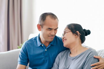 Portrait of Asian mature couple sitting in the living room. wife and husband hugging, holding hand with happiness and cheerful. Love, safety, and insurance family concept.