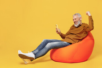 Full size body length cheery elderly gray-haired man 40s years old in brown shirt sit in bag chair hold use mobile cell phone doing winner gesture isolated on plain yellow background studio portrait.