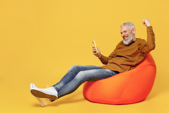 Full Size Body Length Cheery Elderly Gray-haired Man 40s Years Old In Brown Shirt Sit In Bag Chair Hold Use Mobile Cell Phone Doing Winner Gesture Isolated On Plain Yellow Background Studio Portrait.