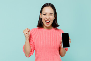 Young happy woman of Asian ethnicity 20s wear pink sweater hold in hand use mobile cell phone with blank screen workspace area do winner gesture isolated on pastel plain light blue background studio.