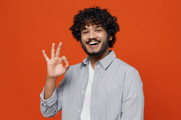Fototapeta premium Cheerful exultant happy overjoyed young bearded Indian man 20s years old wear blue shirt showing okay ok gesture isolated on plain orange background studio portrait. People emotions lifestyle concept