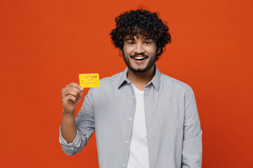 Cheerful smiling fun happy fascinating charming young bearded Indian man 20s years old blue shirt hold in hand credit bank card looking camera isolated on plain orange background studio portrait