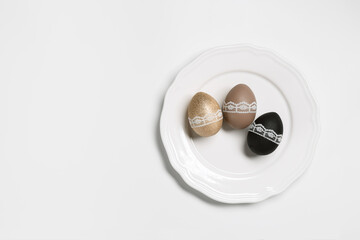 Easter eggs golden, black and beige colored in white plate on white background. Symbol Easter holiday, dyed chicken egg bright trendy colors and decorated lace tape. Top view