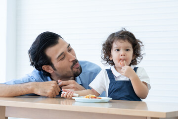 Obraz na płótnie Canvas Caucasian young father with beard looking and cheer up little cute daughter trying to eat spaghetti with spoon by herself at home and adorable kid girl enjoy eating with face is mess up with ketchup