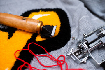 The tufting gun lies on a gray burlap, ready to work with the red yarn in the needle and the...