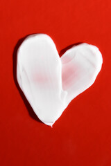Strokes of white color cream in the shape of a heart on a red background, top view. Cosmetic for...