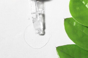 Transparent gel pouring onto a white surface from a pipette, top view. Essence of cosmetic product with green leaves of the plant, body care.