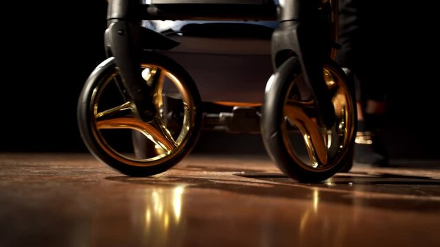 Unrecognizable woman pushes baby pram. Lower part of a child vehicle with shining details. Dark backdrop.
