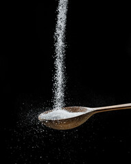 White refined sugar crystals falling down into the wooden spoon at black background.