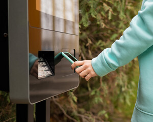 A woman uses a street self-service terminal for contactless payment with a smartphone.