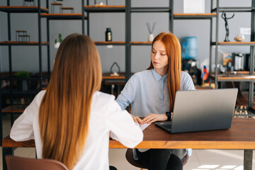 View from back to female applicant during job interview talking with young woman employers sitting at desk opposite each other. Businesswoman sitting at desk interviewing lady candidate in office.