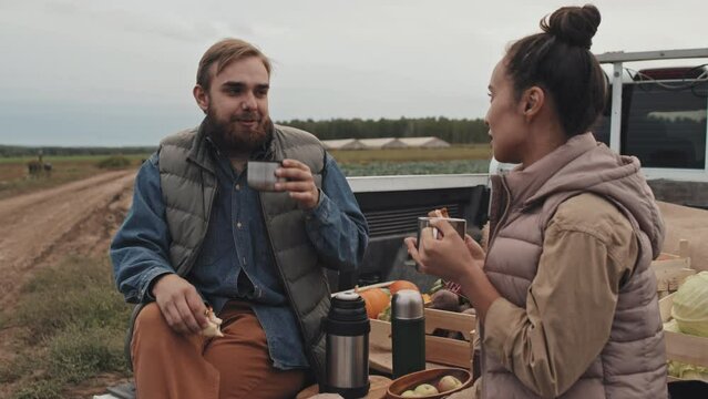 Young adult man and woman sitting relaxed on pickup truck cargo having lunch break during workday on farm field