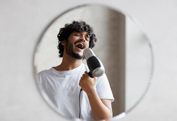 Joyful indian man having fun in the morning in bathroom, holding hairdryer and singing as in...