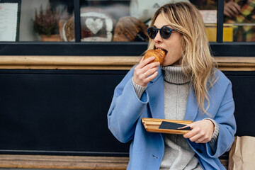 Hungry blonde woman, taking a bite from the croissant, while looking towards the street.