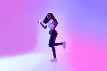 Fototapeta na wymiar Cheerful African American female with smartphone running in neon light, showing mockup for mobile app or website design