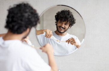 Indian man cleaning teeth in the morning, applying toothpaste on toothbrush, looking at mirror in bathroom, free space