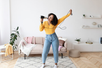 Positive young African American woman using hair dryer as microphone, singing and dancing, having...